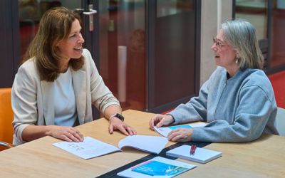 Twee vrouwen zitten aan tafel in gesprek, met documenten en brochures voor zich, terwijl ze elkaar glimlachend aankijken.
