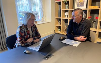 Twee vrouwen zitten aan tafel voor een gesprek, met papieren en een brochure op tafel, in een moderne kantooromgeving.